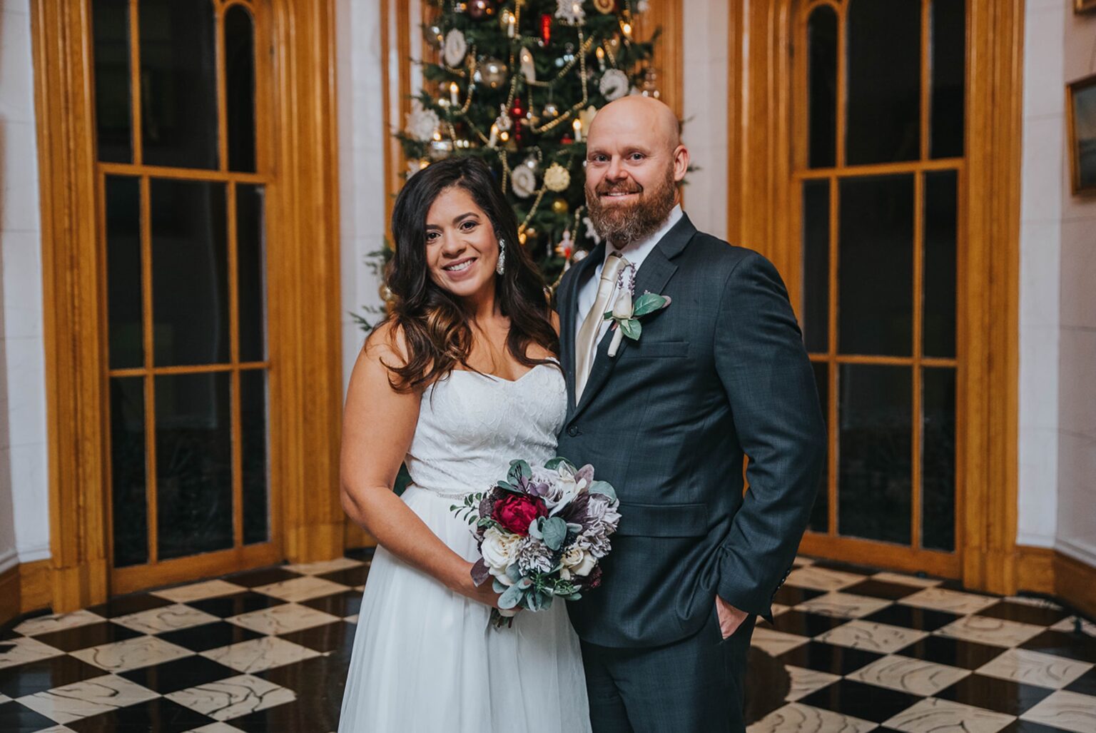 Elopement Wedding A smiling bride in a white dress holds a bouquet of flowers while standing next to a groom in a dark suit and tie. They are in front of a decorated Christmas tree and wooden framed windows in a venue with checkered black and white floors. Elopements Inc